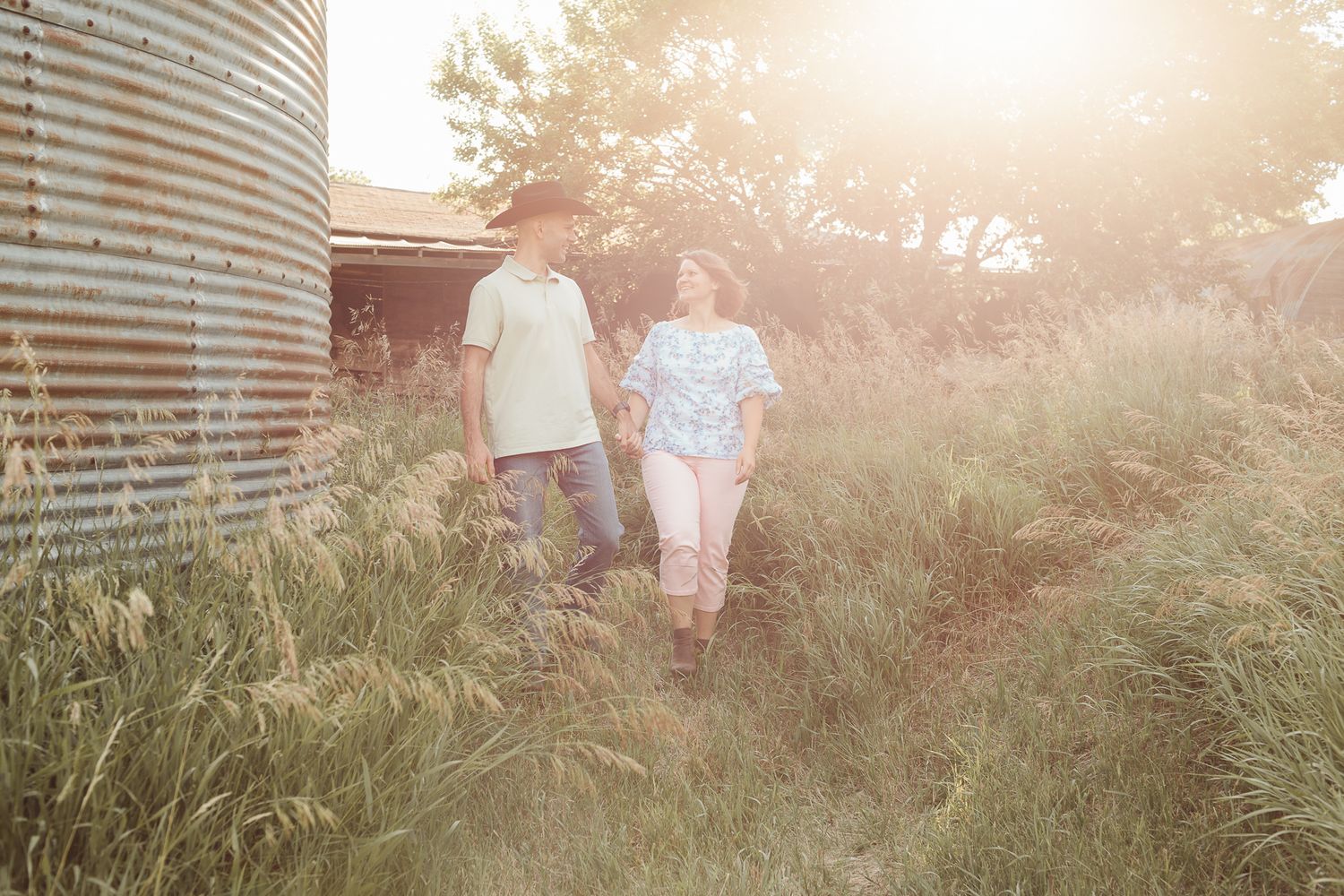 Couple walking hand in hand through tall green grass near grain silo in golden sunset light.