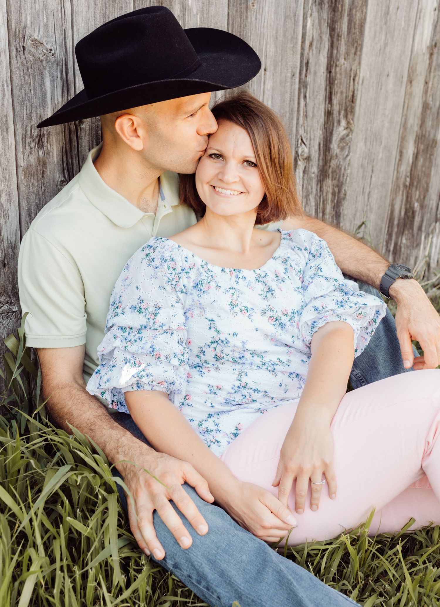 Romantic couple posing by weathered wood fence, man in black cowboy hat kissing woman on cheek outdoors.