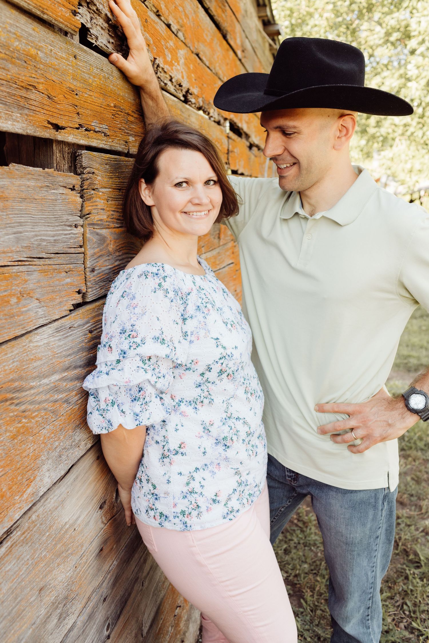 Couple leaning against rustic wooden barn wall, man in black cowboy hat smiling at woman in floral blouse.