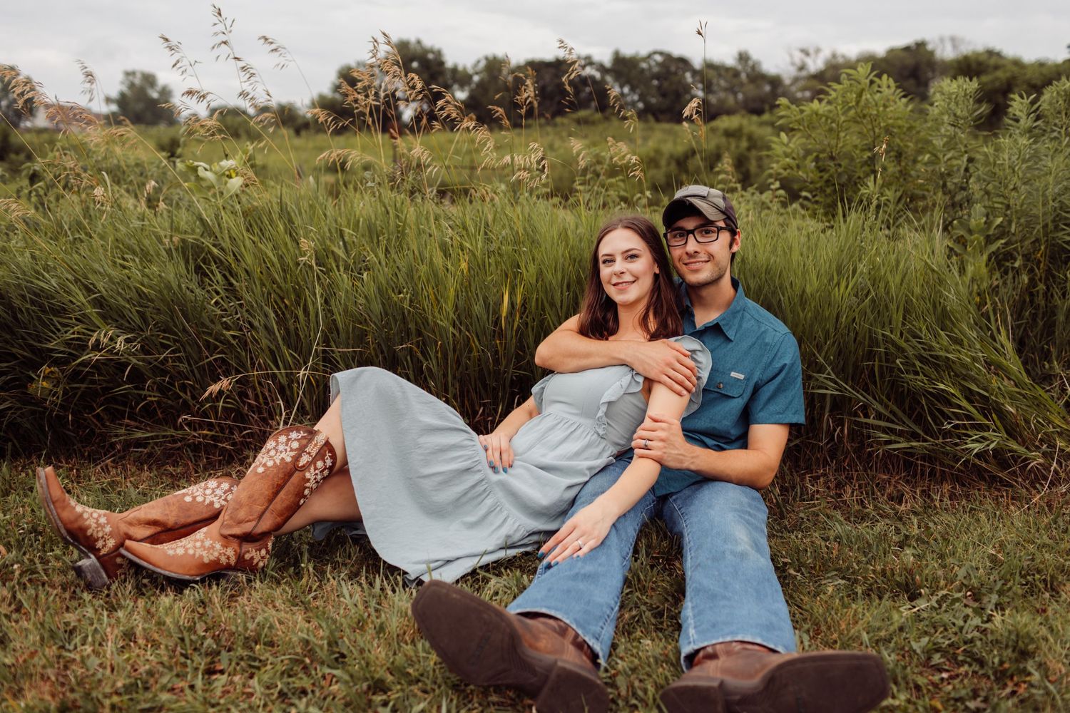 Happy couple sitting together in tall grass field, woman in light blue dress with cowboy boots, casual outdoor portrait.