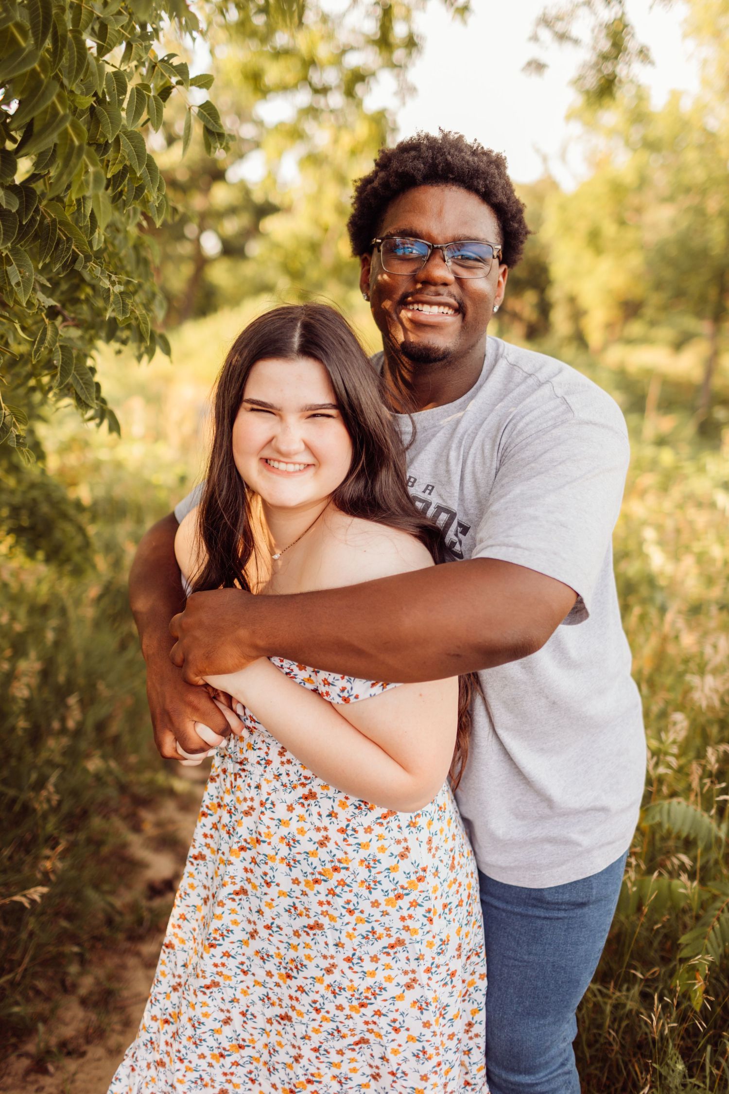 Young man hugging woman from behind while smiling outdoors in golden sunlight.