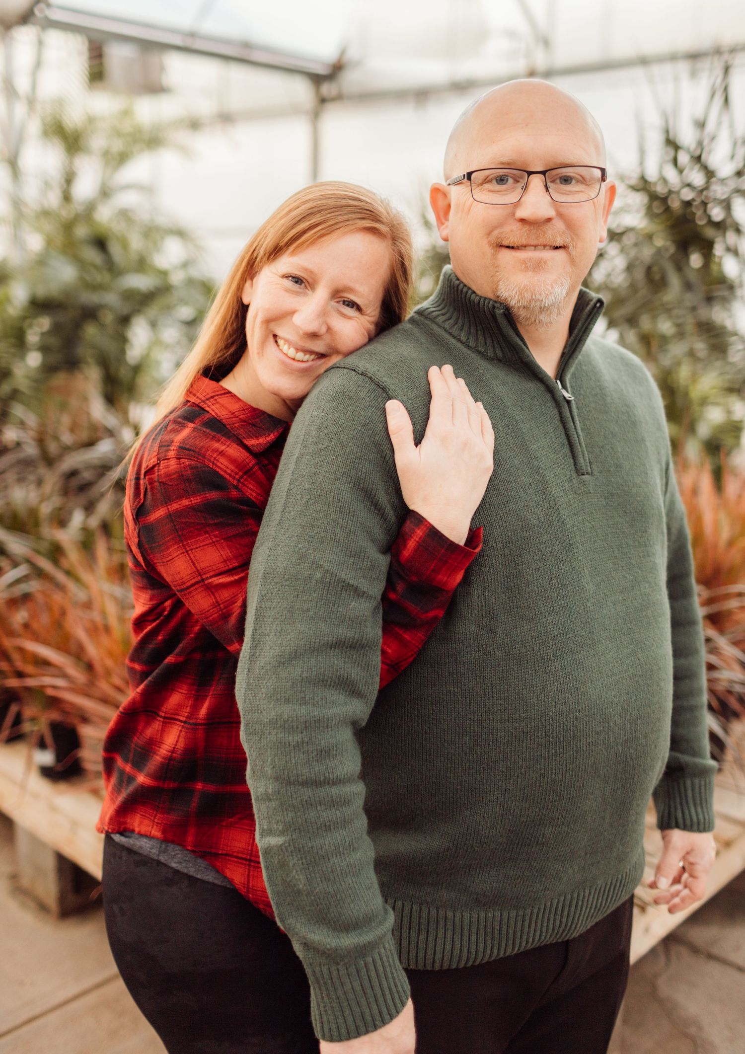 Smiling redhead woman leaning on bald man in green sweater inside a lush greenhouse setting.