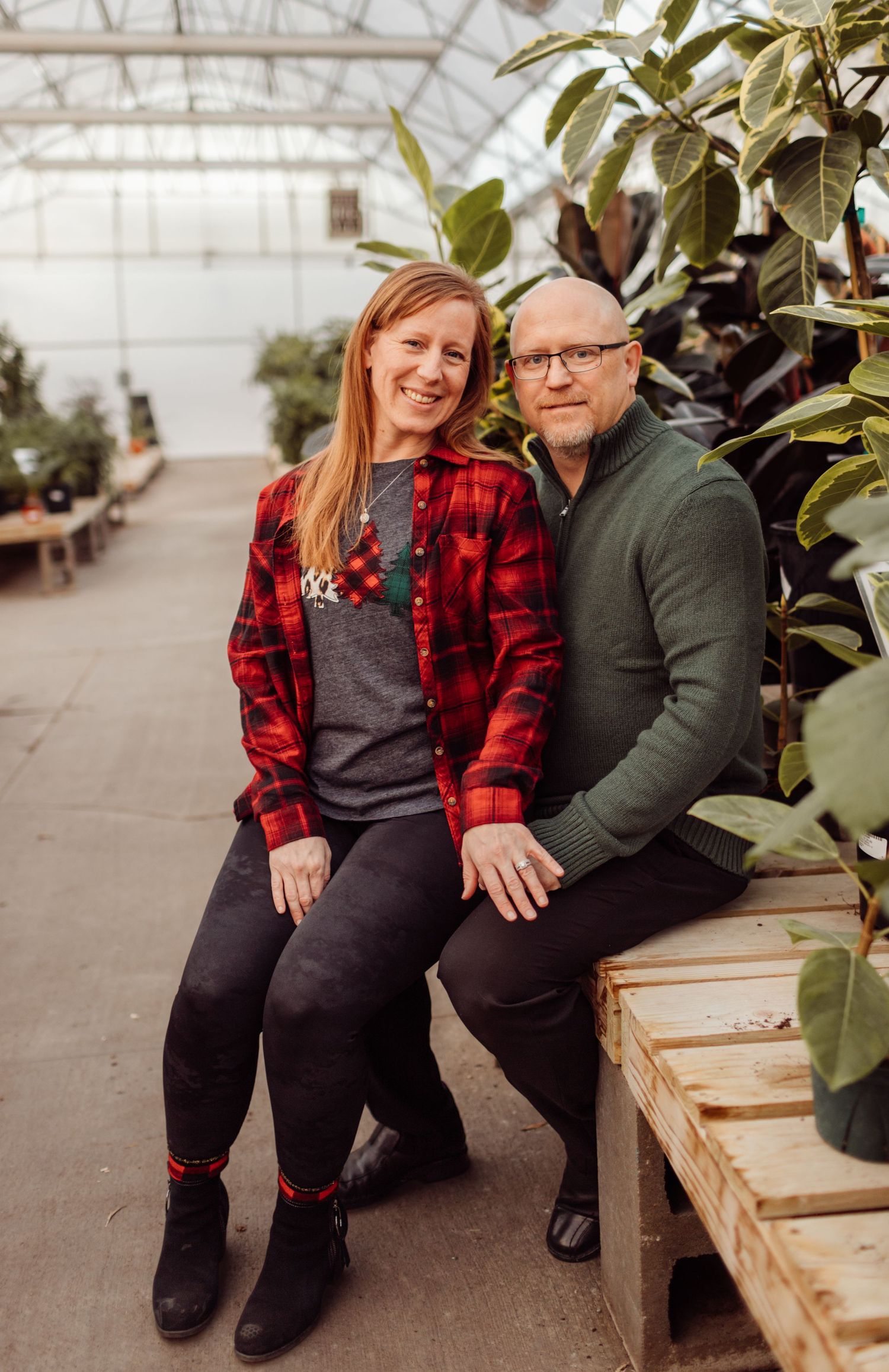 Couple sitting together among tropical plants in a greenhouse, both smiling warmly at the camera.