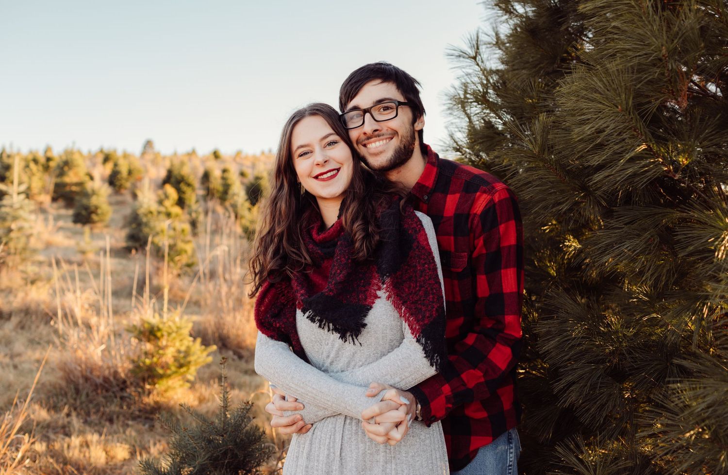 Happy couple embracing near evergreen tree in golden field wearing cozy plaid winter attire.