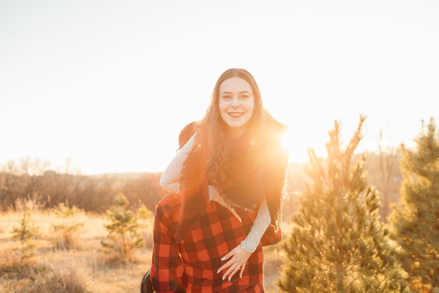 Woman smiling warmly with sun flare behind her at an outdoor Christmas tree farm.