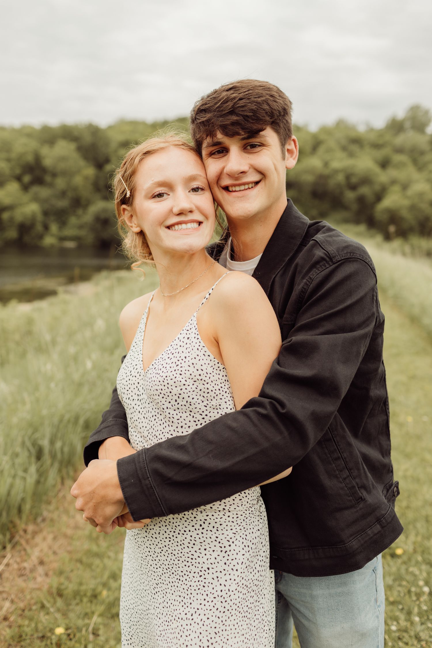 Happy young couple embracing outdoors near a river, man hugging woman from behind in a green field.