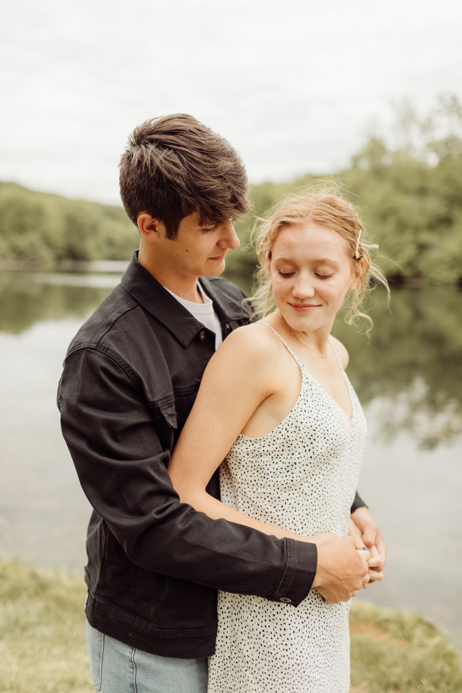 Young couple embracing tenderly by a serene lake, man in black jacket holding woman in floral dress.
