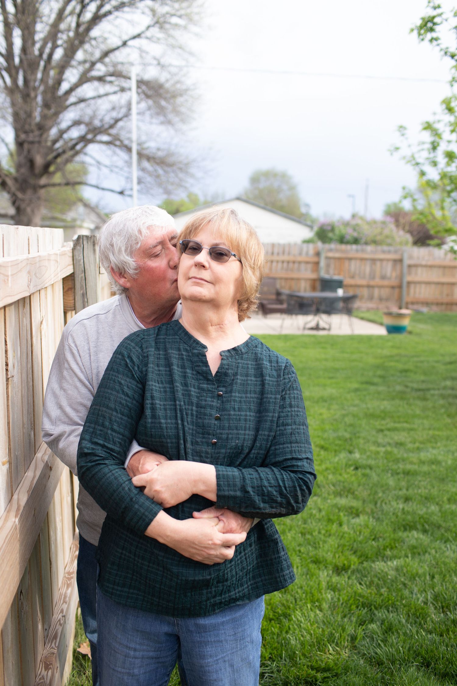 Senior man kissing woman on cheek in a backyard garden, woman smiling contentedly by wooden fence.
