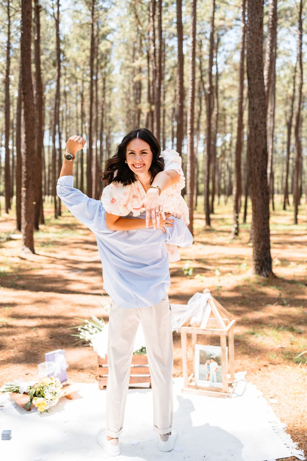 A joyful woman celebrates outdoors at a forest picnic, raising her arms while holding a champagne bottle.
