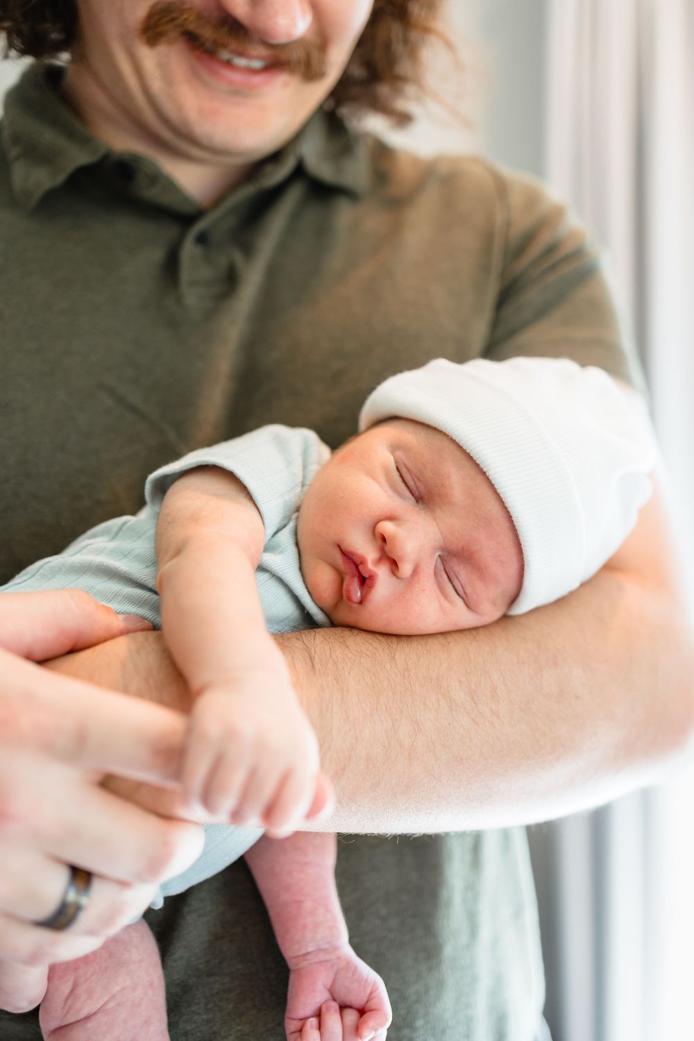 A smiling father with a mustache gently cradles his sleeping newborn baby wearing a white hat.