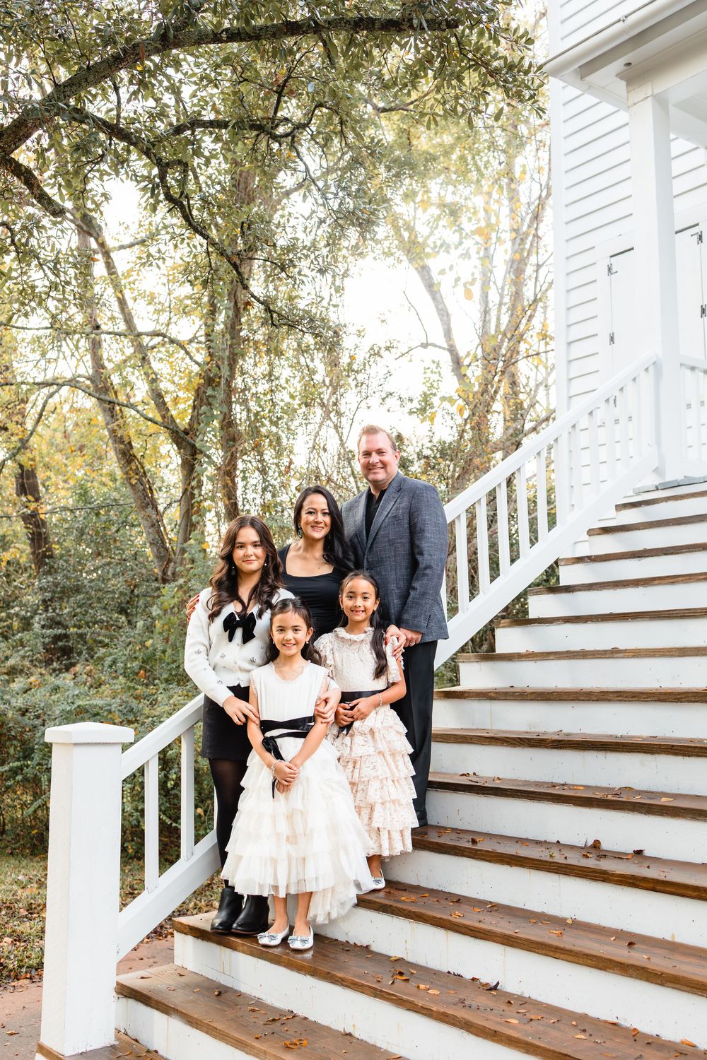 A family of five poses on outdoor wooden stairs, with two parents and three daughters in elegant dresses.