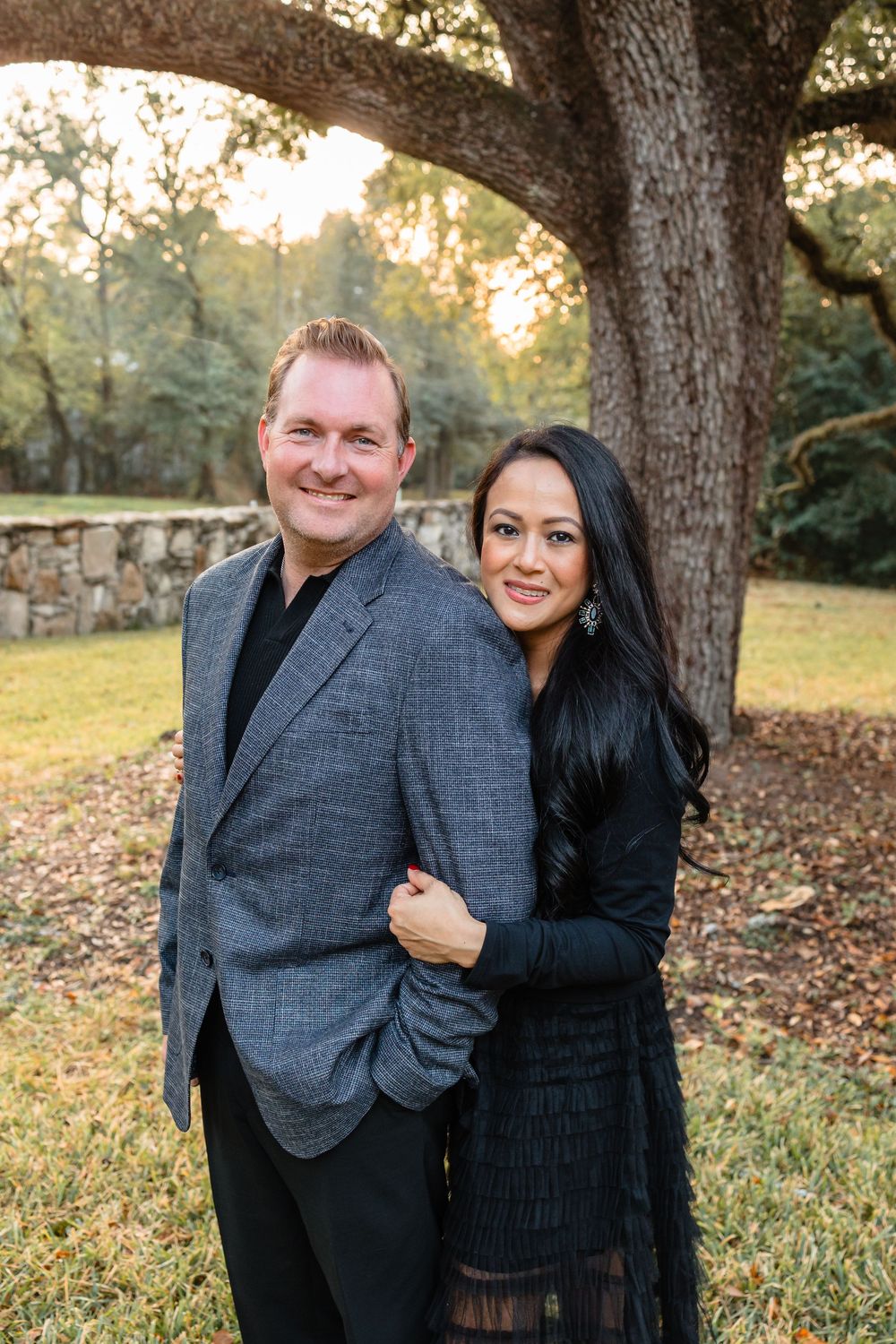 A smiling couple dressed in elegant black attire poses together under a large oak tree at dusk.