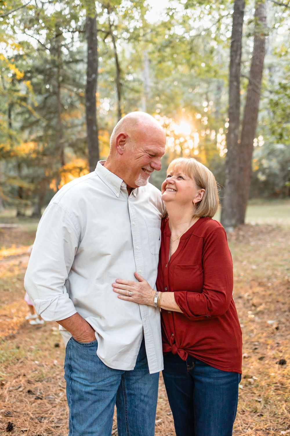 A happy older couple shares a warm smile while embracing in a sunlit autumn forest setting.