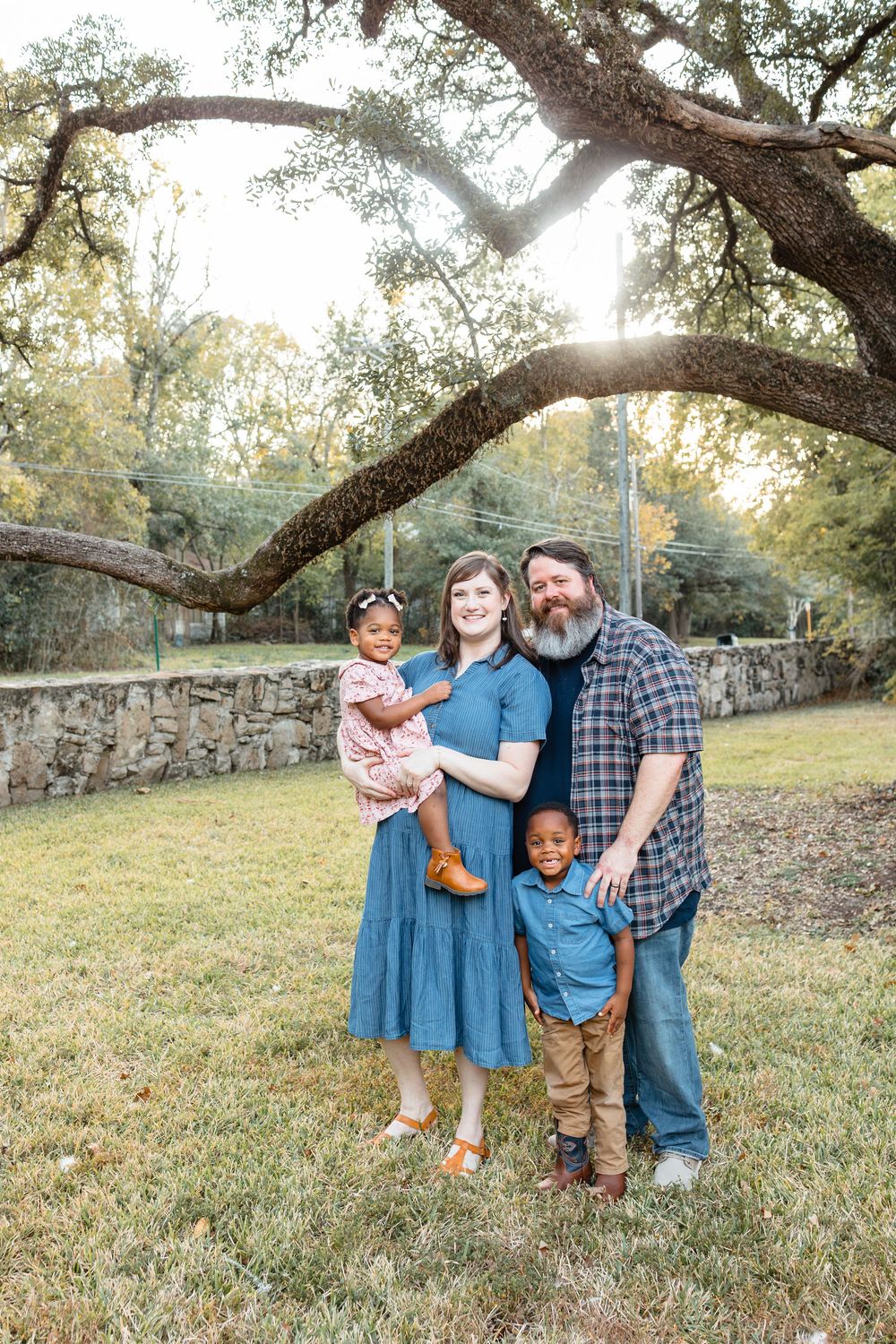 A bearded father and mother pose outdoors with their two young adopted children under a large oak tree.