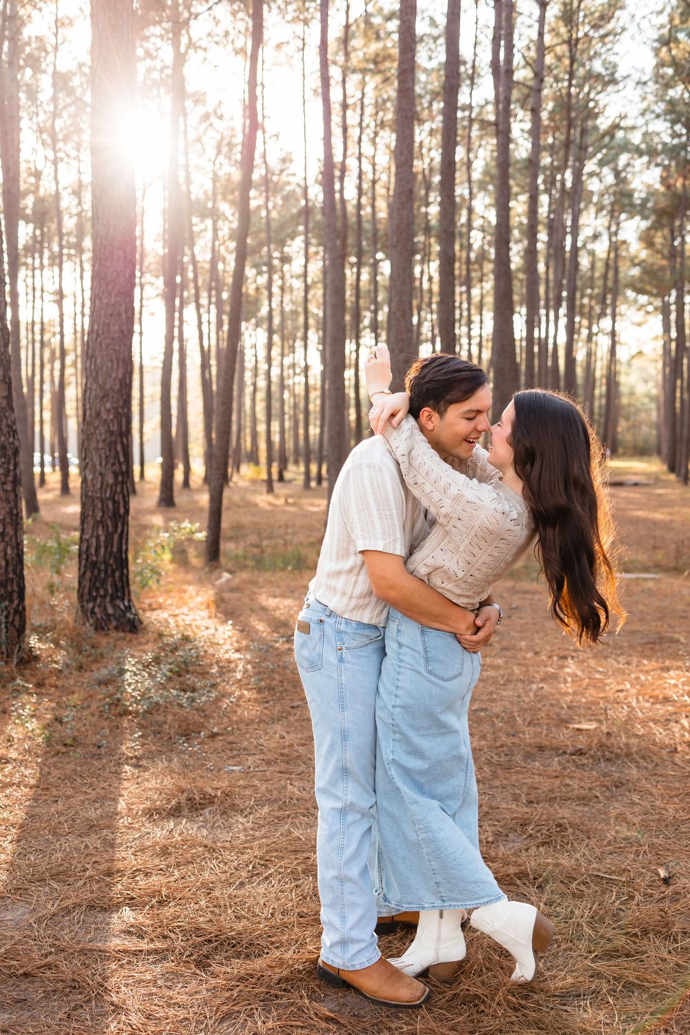 Couple embracing and smiling at each other in a sunlit pine forest during golden hour.