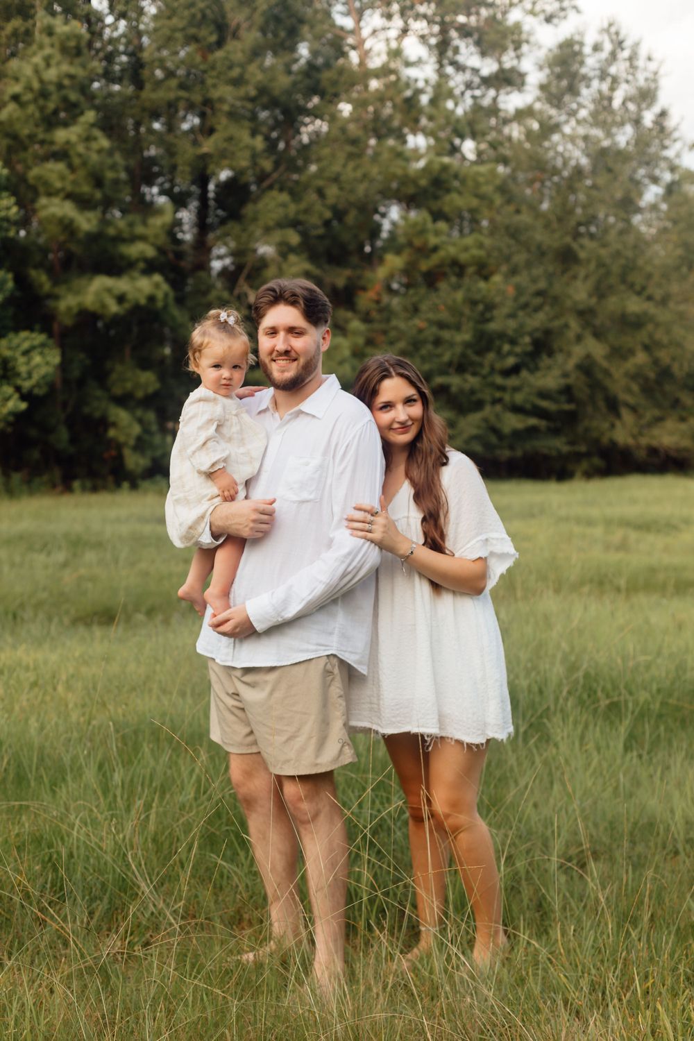 Happy young family with a toddler posing barefoot in a lush green meadow surrounded by trees.