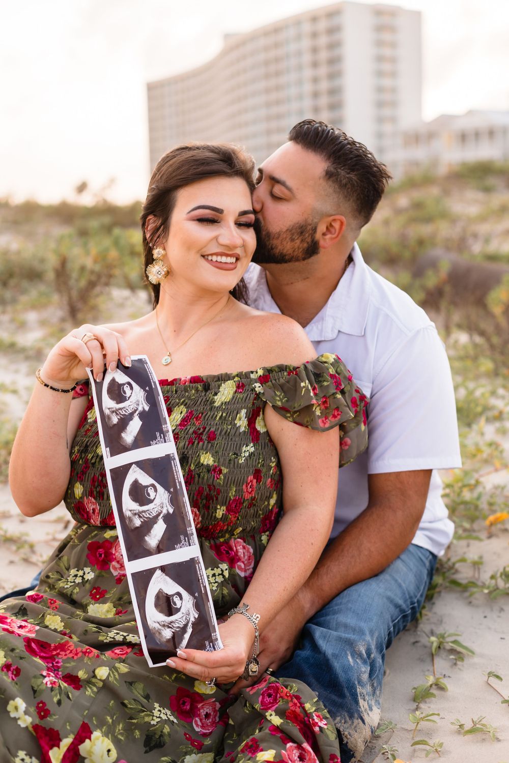 Couple at the beach holding ultrasound photos, announcing pregnancy with a kiss on sandy dunes.