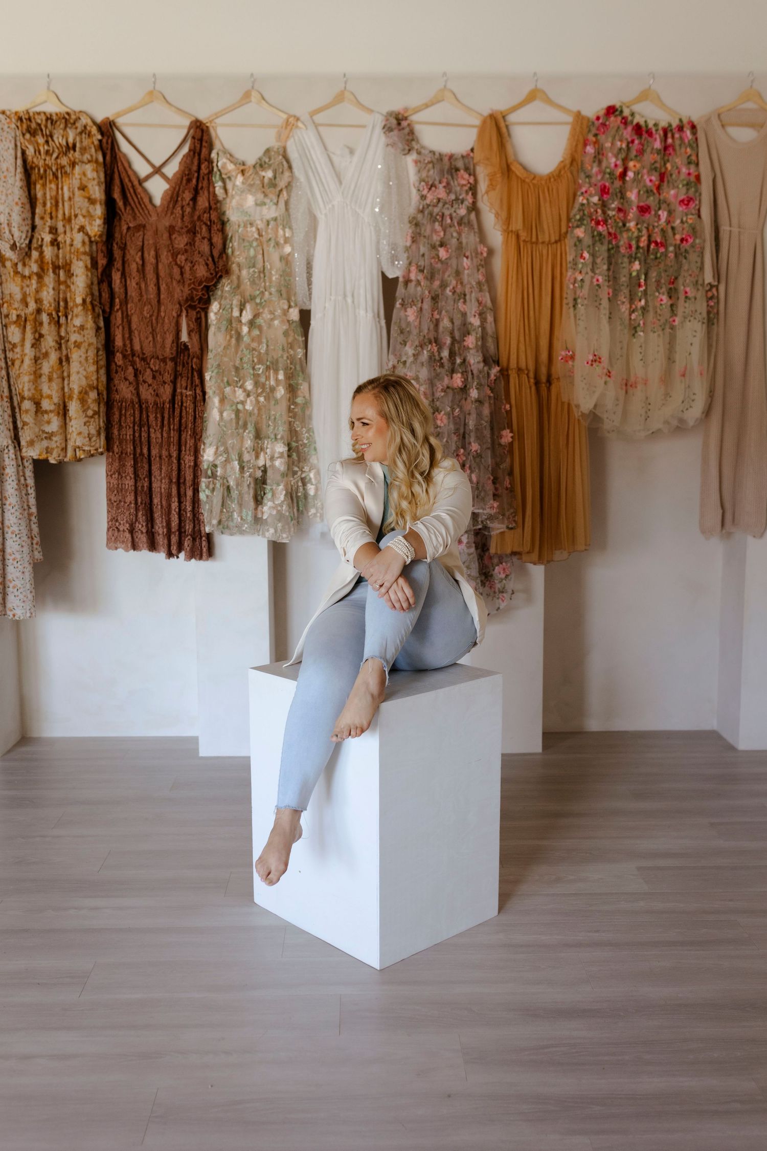 Blonde woman sitting on white pedestal in front of hanging dresses display.