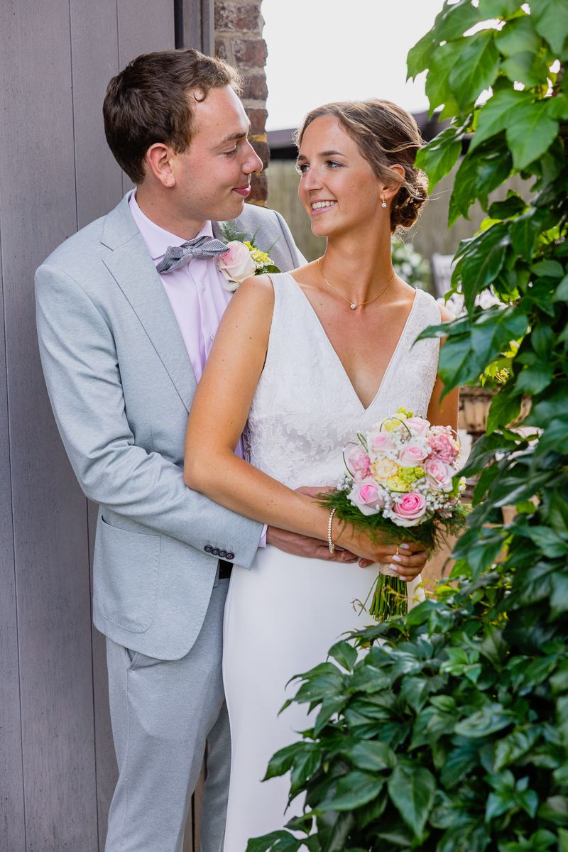 A romantic wedding couple in light gray suit and white dress share a tender moment among green foliage.