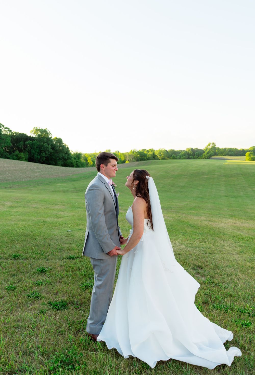 Bride and groom holding hands in a lush green field at golden hour, wearing white gown and gray suit.