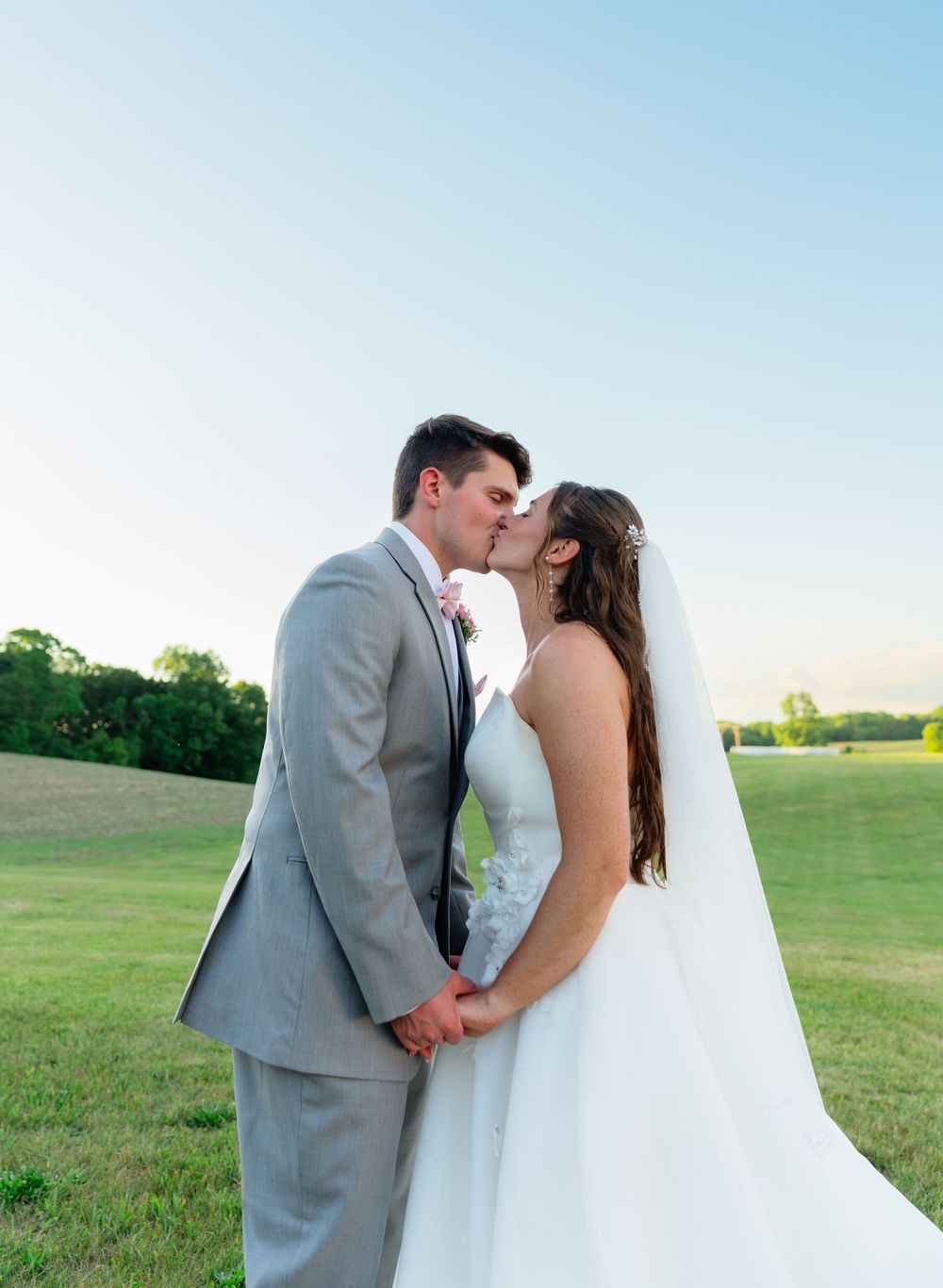 A bride and groom share a romantic kiss outdoors on their wedding day, with a green field and clear blue sky behind them.