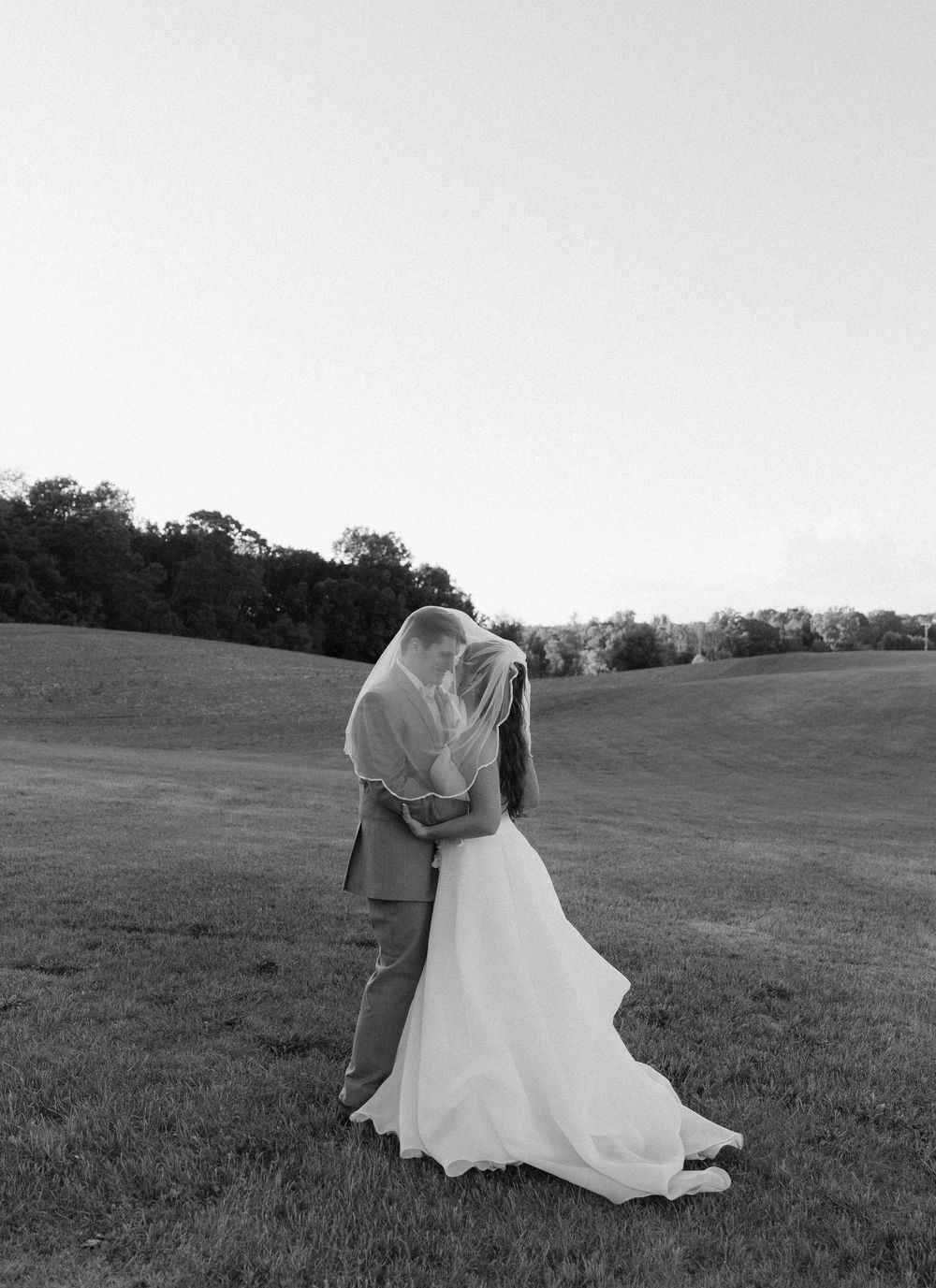 Black and white photo of bride and groom sharing a kiss under veil in an open field on wedding day.