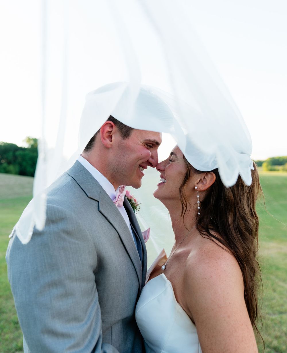 A bride and groom share a romantic moment under a flowing white veil outdoors on their wedding day.