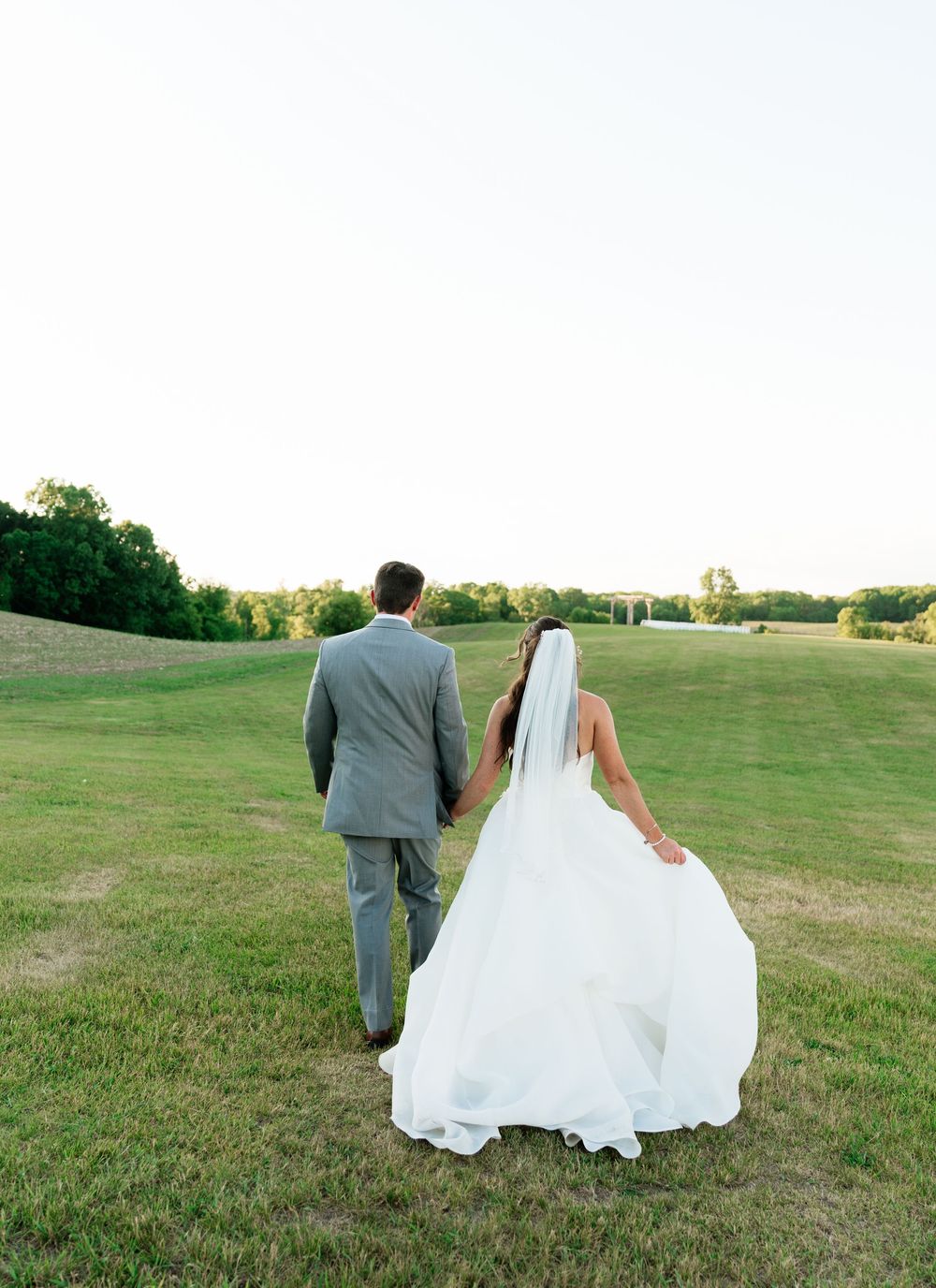 Bride and groom walking hand in hand across a lush green field in wedding attire, white gown trailing behind.