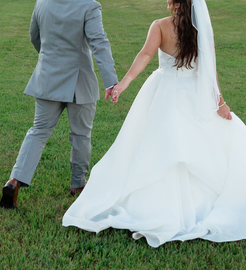A bride in a white wedding gown and groom in a gray suit walk hand in hand across a lush green lawn.