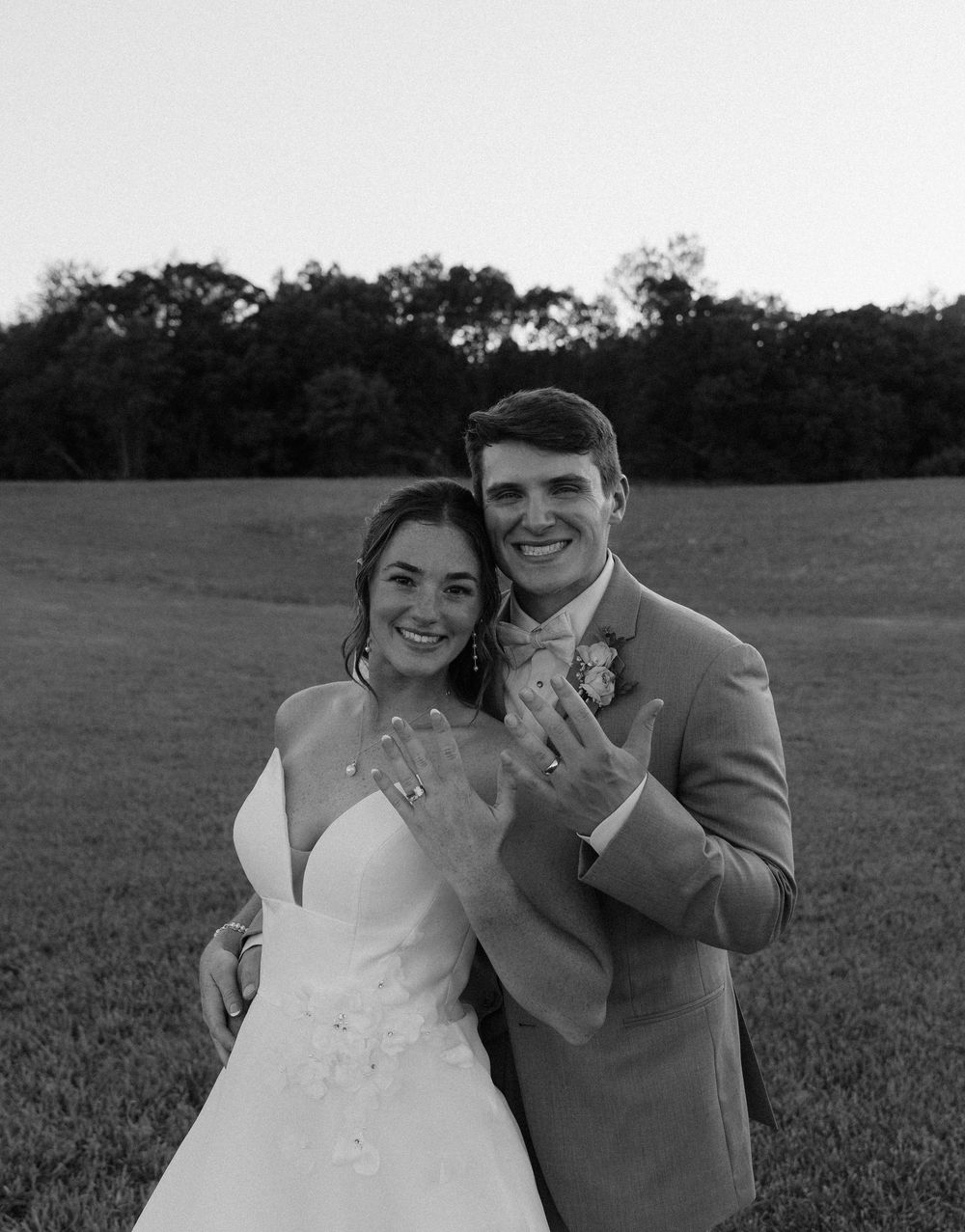Black and white photo of newlyweds showing off wedding rings in a field, bride in gown and groom in suit smiling.