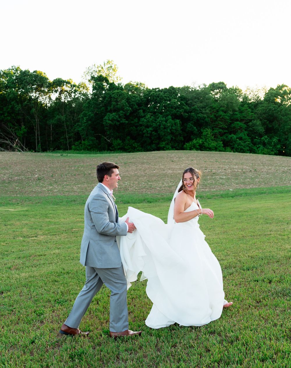 Groom in gray suit helps bride carry her white wedding gown while walking together across a green field.