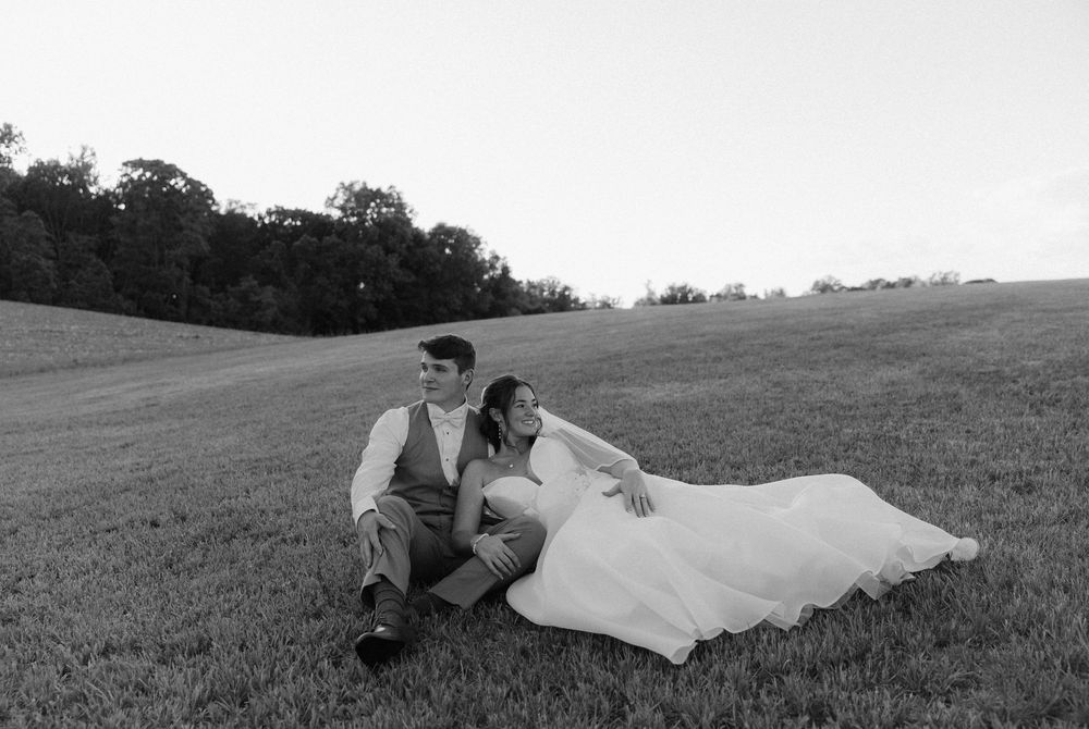 Black and white photo of a bride and groom sitting together in a lush green field on their wedding day.