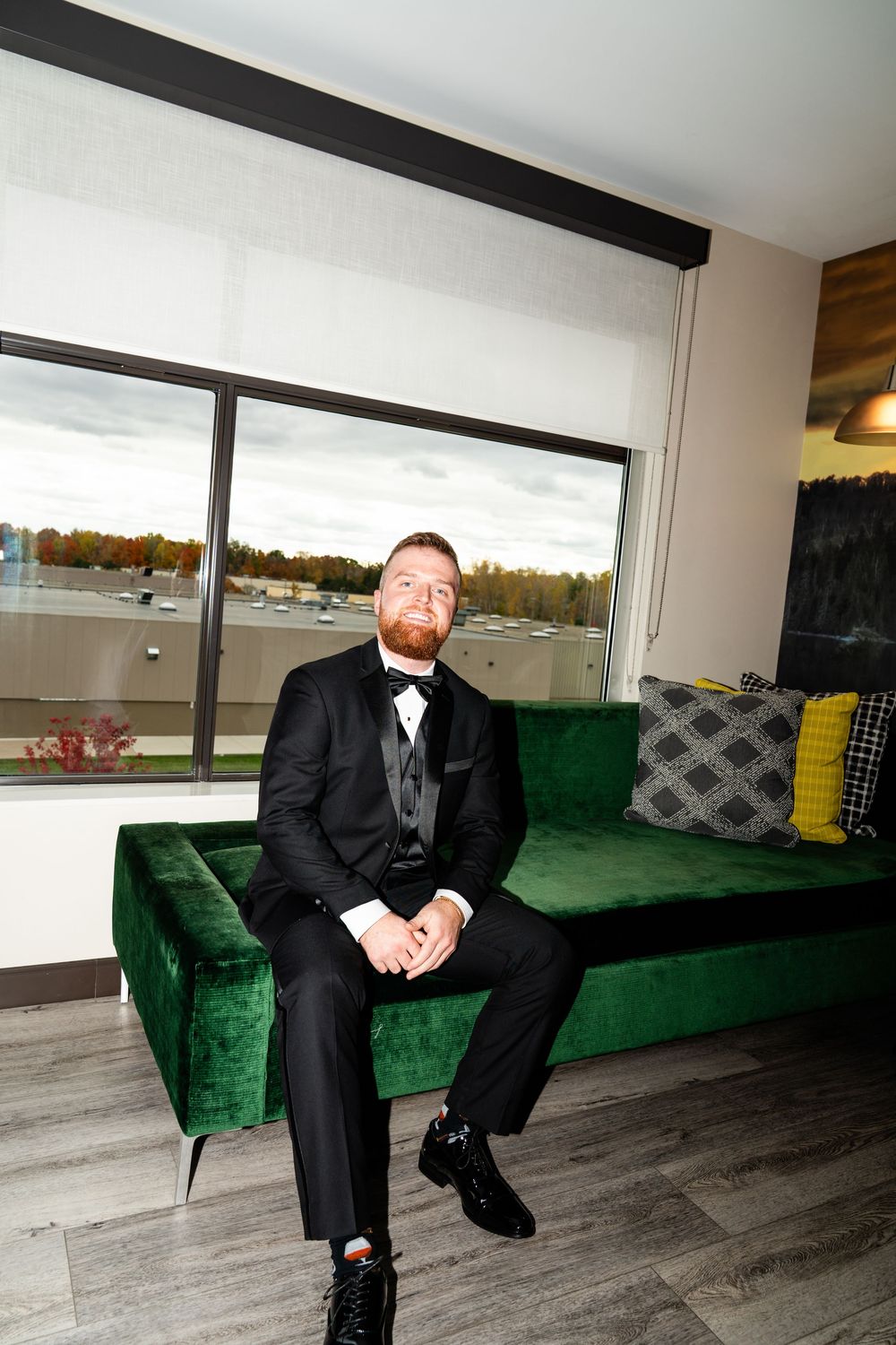 Groom seated on emerald green velvet couch in formal wedding attire.