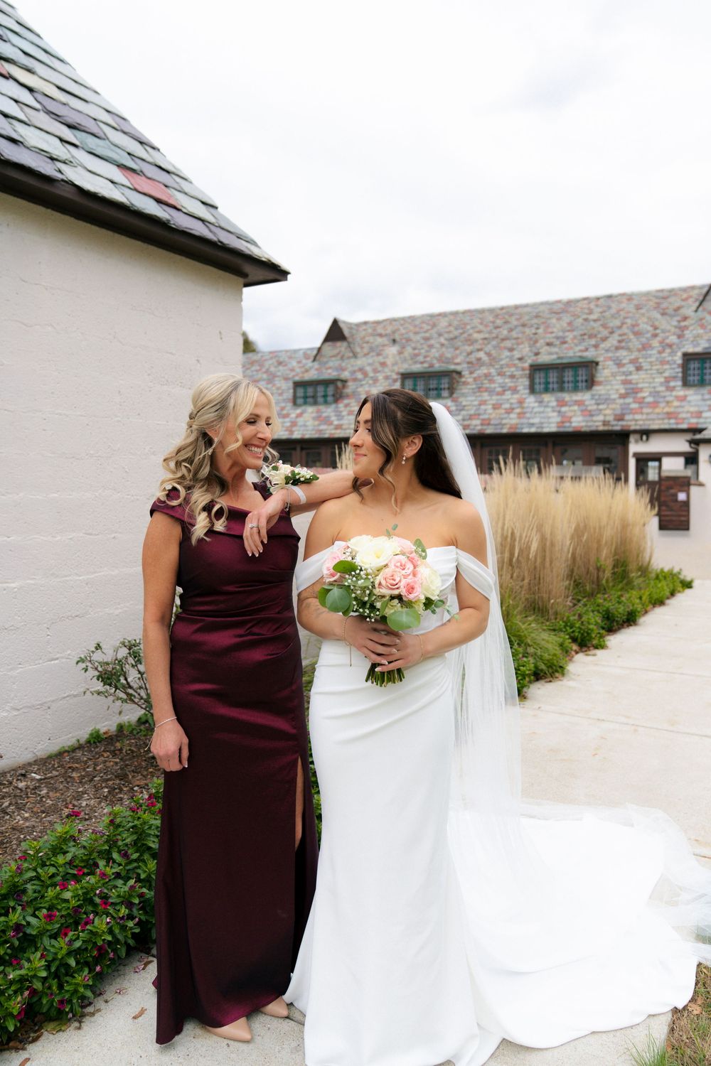 Bride in white gown with bridesmaid in burgundy dress walking along stone pathway.