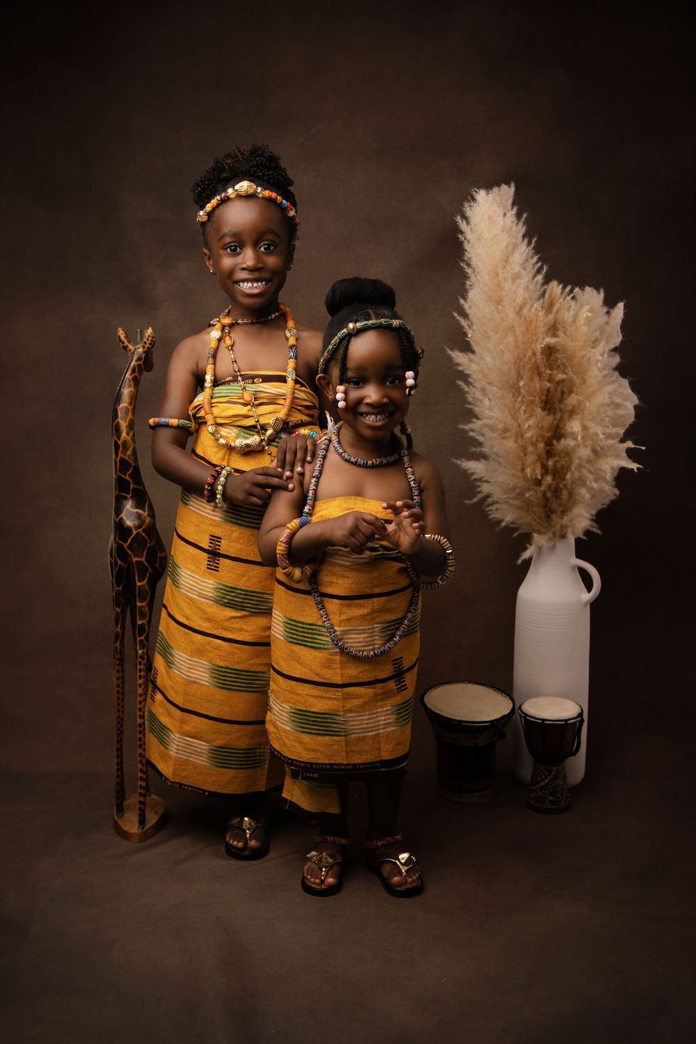 Two children wearing matching yellow and brown African print dresses pose together in a studio setting with pampas grass decor.