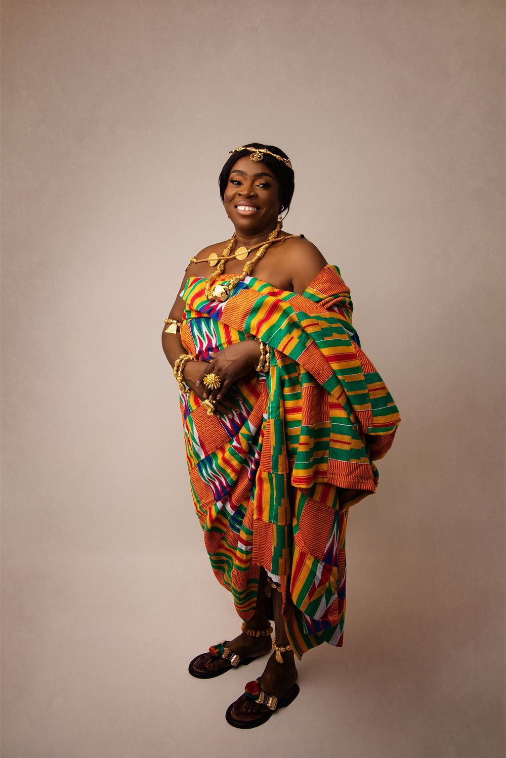 A figure wearing vibrant kente cloth in orange, green and yellow patterns poses against a neutral studio background.