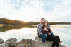 Couple sits together on rocky shore beside tranquil lake during golden sunset with autumn trees in background.