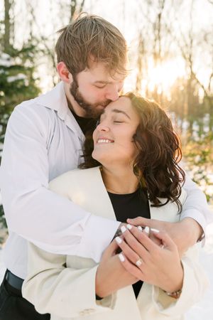 A romantic couple embraces outdoors during golden hour sunlight in a winter forest setting.