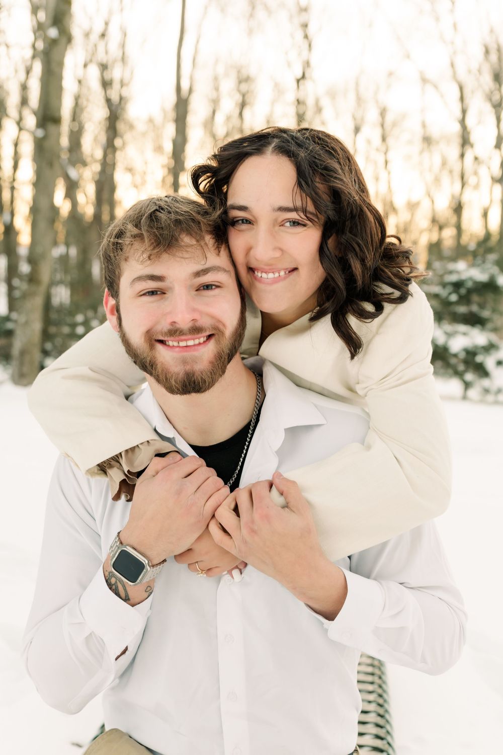A couple wearing white winter outfits share intimate moments in a snowy forest setting.
