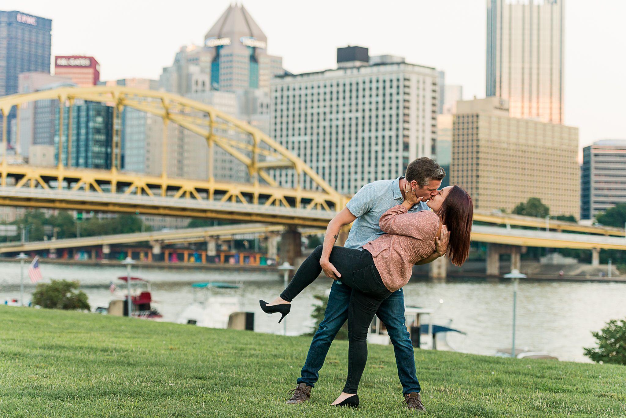 A couple shares a romantic dip pose by the river with Pittsburgh's iconic yellow bridge and city skyline in the background.