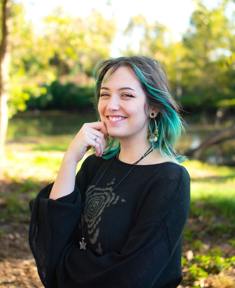 Portrait of someone wearing a black sweater with teal hair in a sunny outdoor setting.