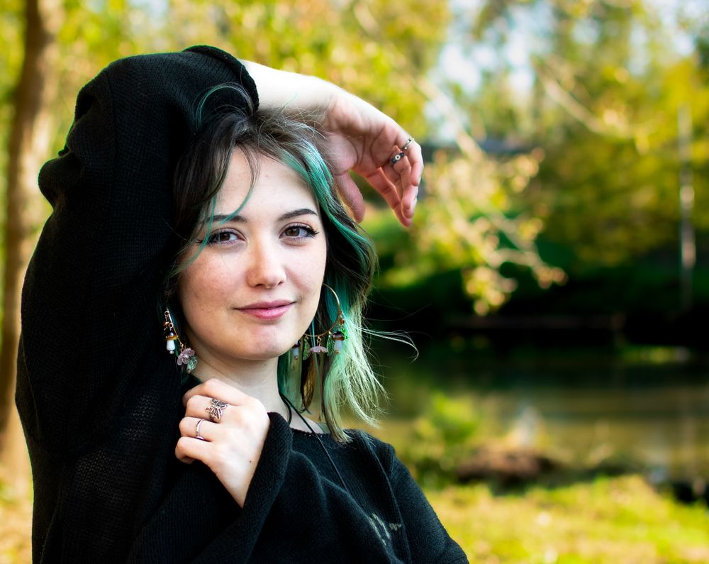 Artistic outdoor portrait with teal-dyed hair and black sweater in golden sunlight.