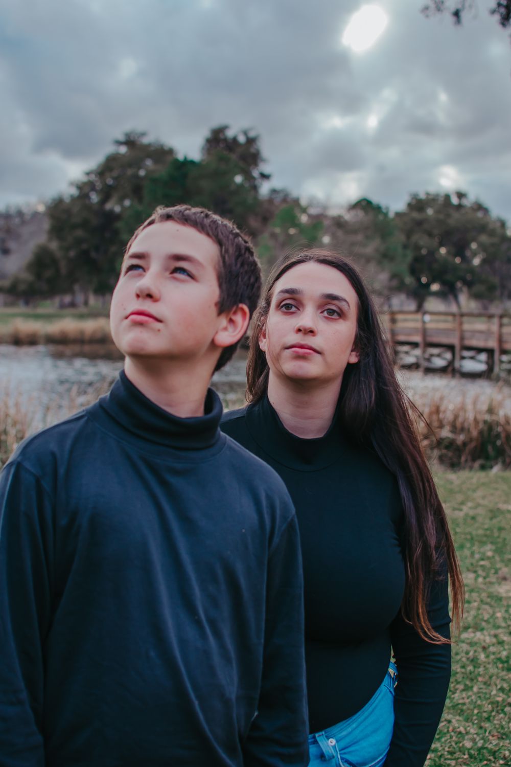Two people in dark sweaters pose together against cloudy sky and water backdrop.
