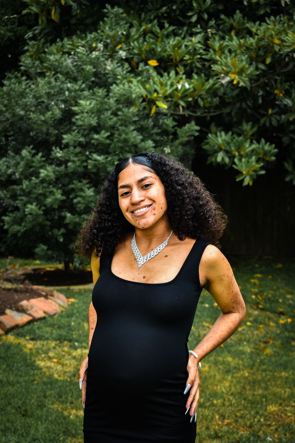 Maternity photoshoot in garden setting with black dress and silver necklace.
