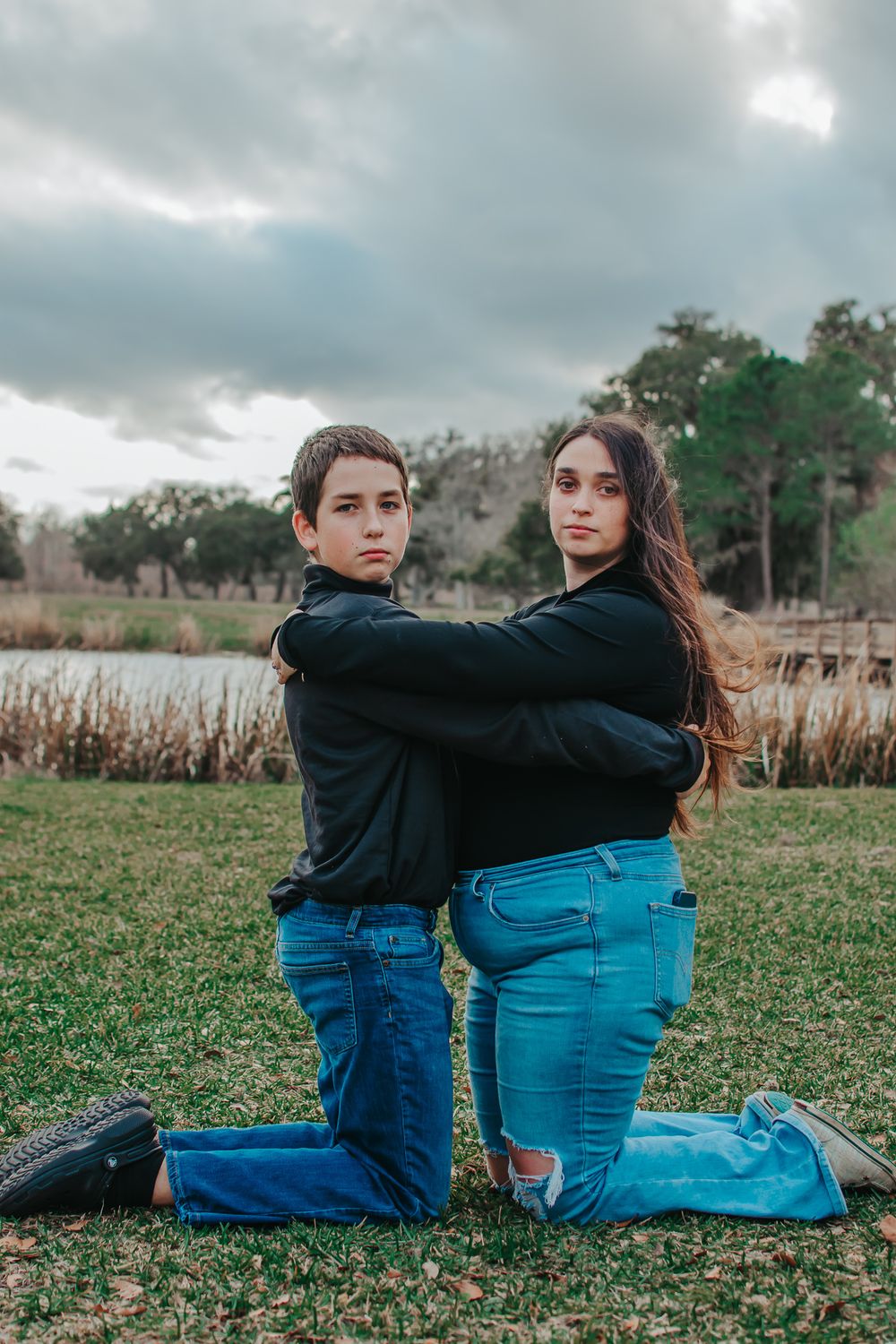 Two people in dark clothing pose on knees in outdoor setting near water.
