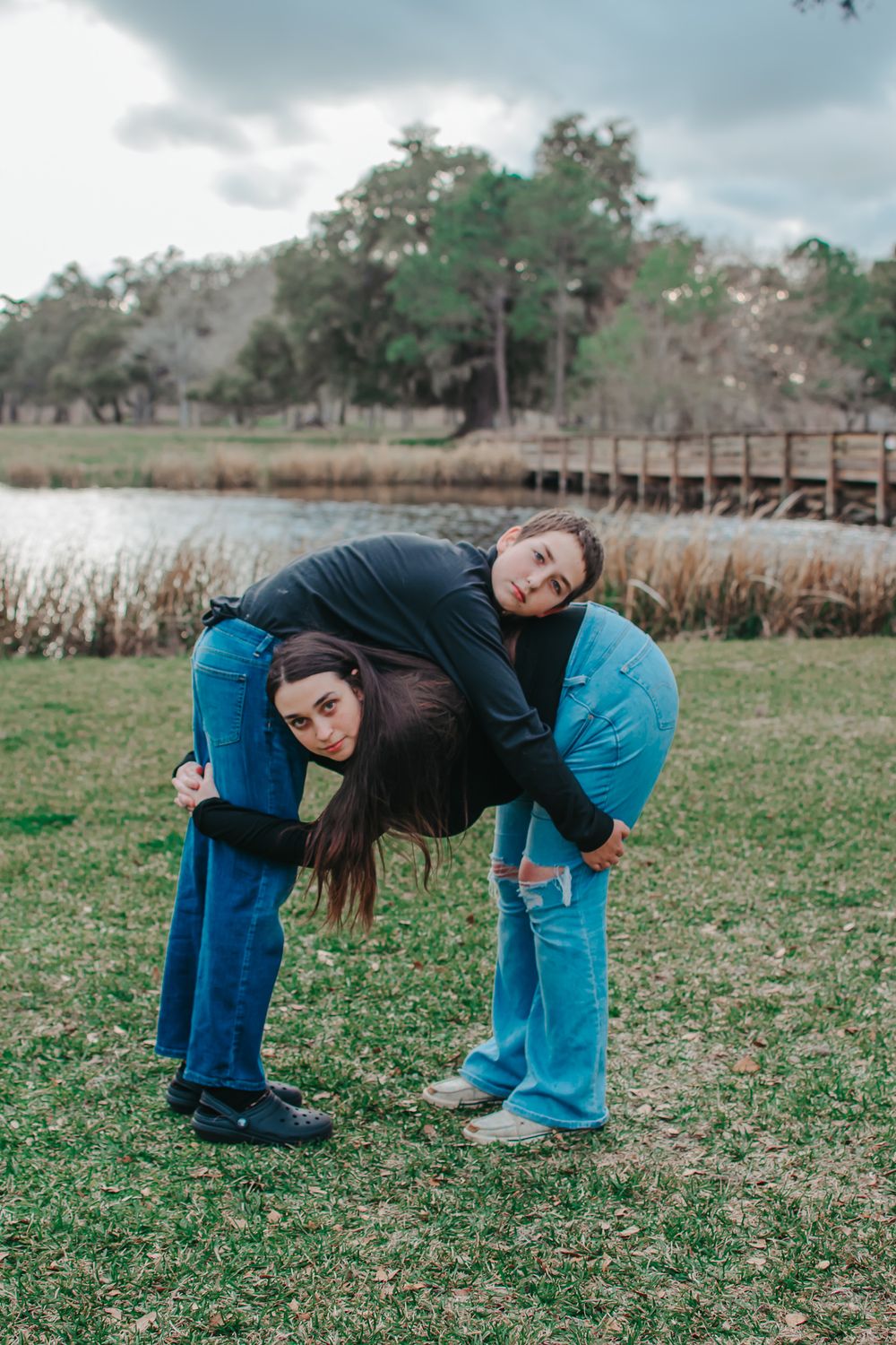 Two people in casual wear pose playfully in park setting with water and boardwalk.