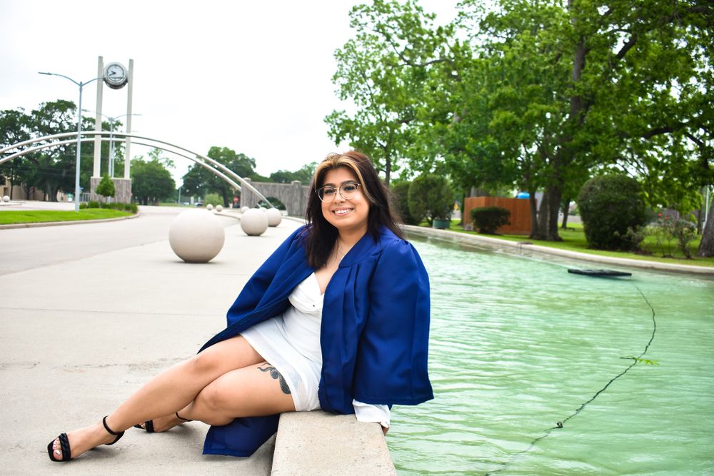 Graduate in blue jacket sits casually by fountain on campus grounds.