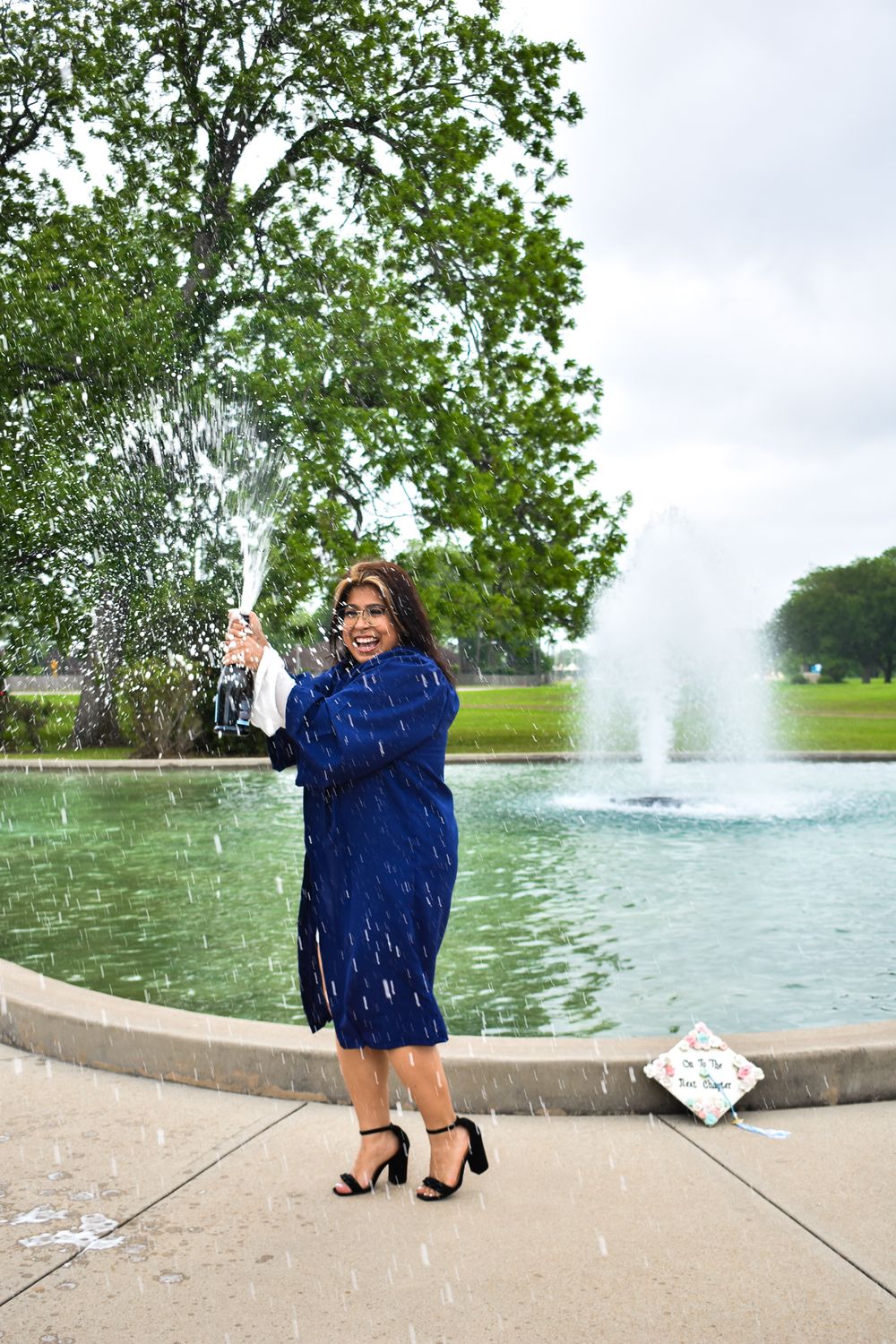 Graduate in blue dress celebrates by fountain with water splashing in background.