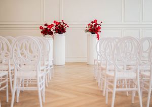 White bentwood chairs arranged in rows with red and white floral arrangements in tall vases for an elegant indoor ceremony.