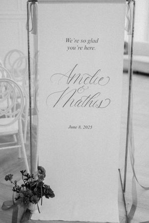 Black and white wedding welcome sign with elegant calligraphy featuring names and date June 8, 2023, with floral decor below.